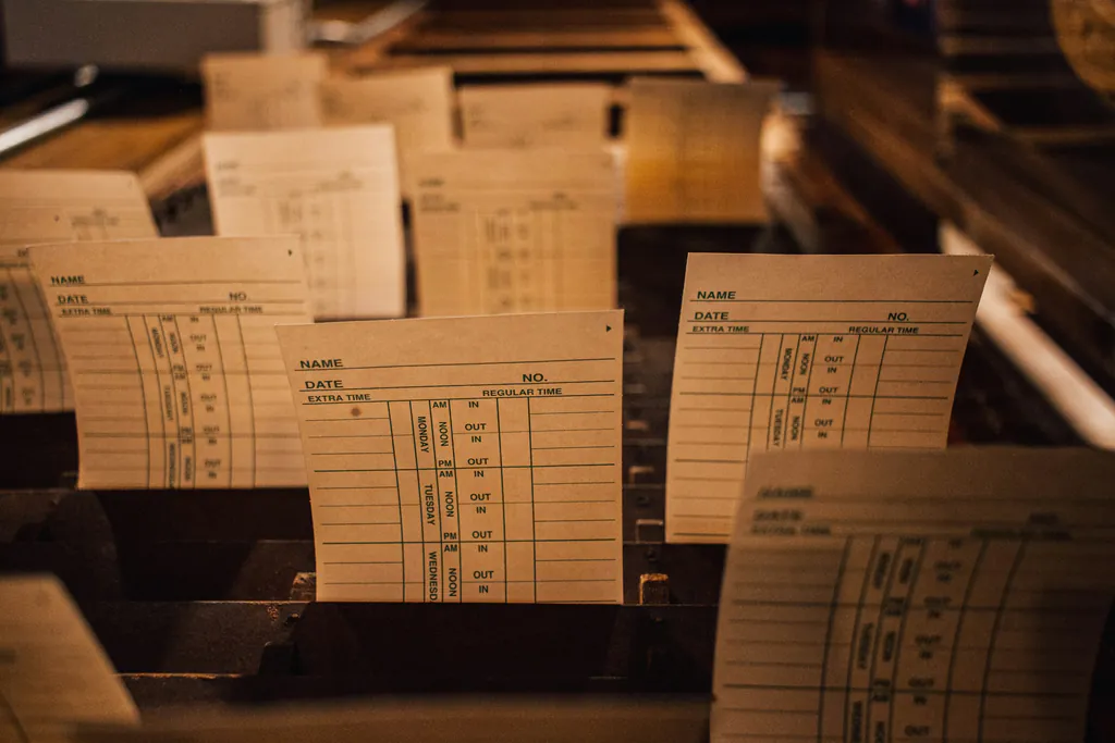 Rows of blank paper time cards with name, date and in and out columns standing in a worn wooden rack