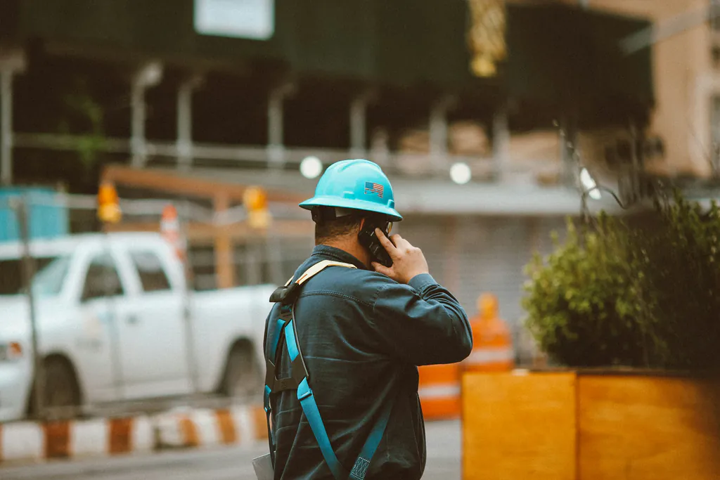 A tradesman in a hard hat and safety harness, seen from behind, holding a phone to their ear on an active job site with a work truck and cones behind them