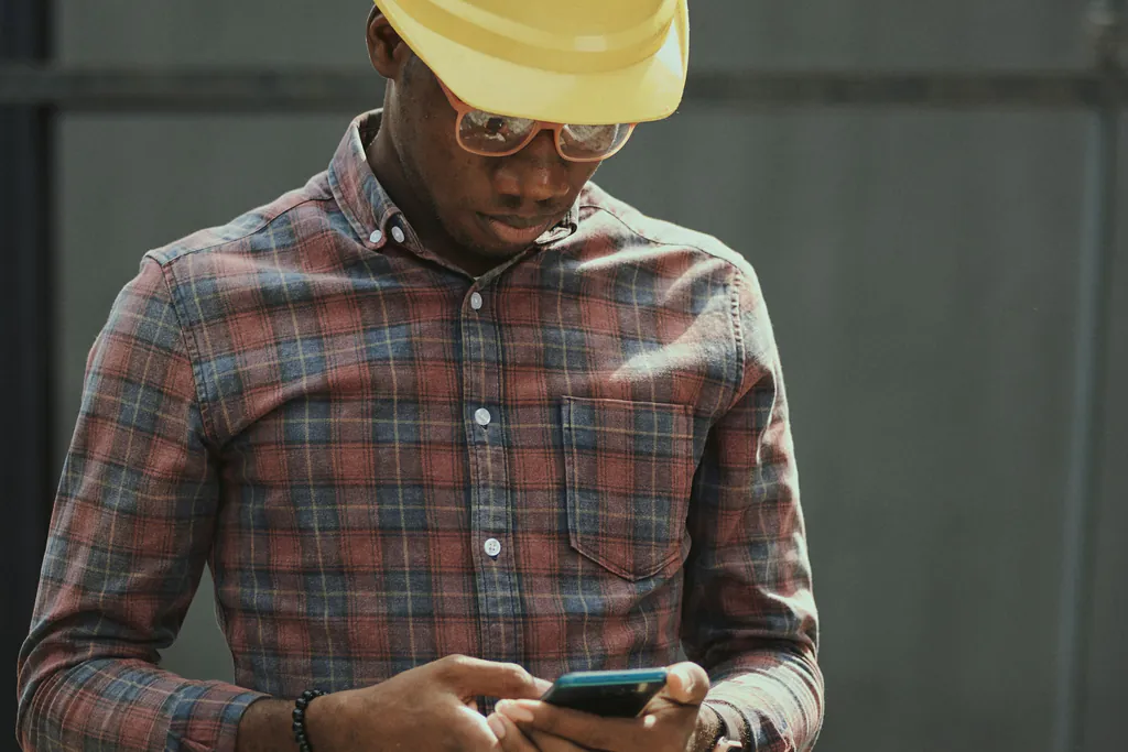An electrician in a hard hat and safety glasses looking down at their phone on site, still waiting to hear back on a quote