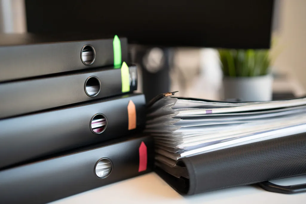 A stack of black ring binders marked with colored flags next to an overstuffed binder of sheet protectors on a project manager's desk