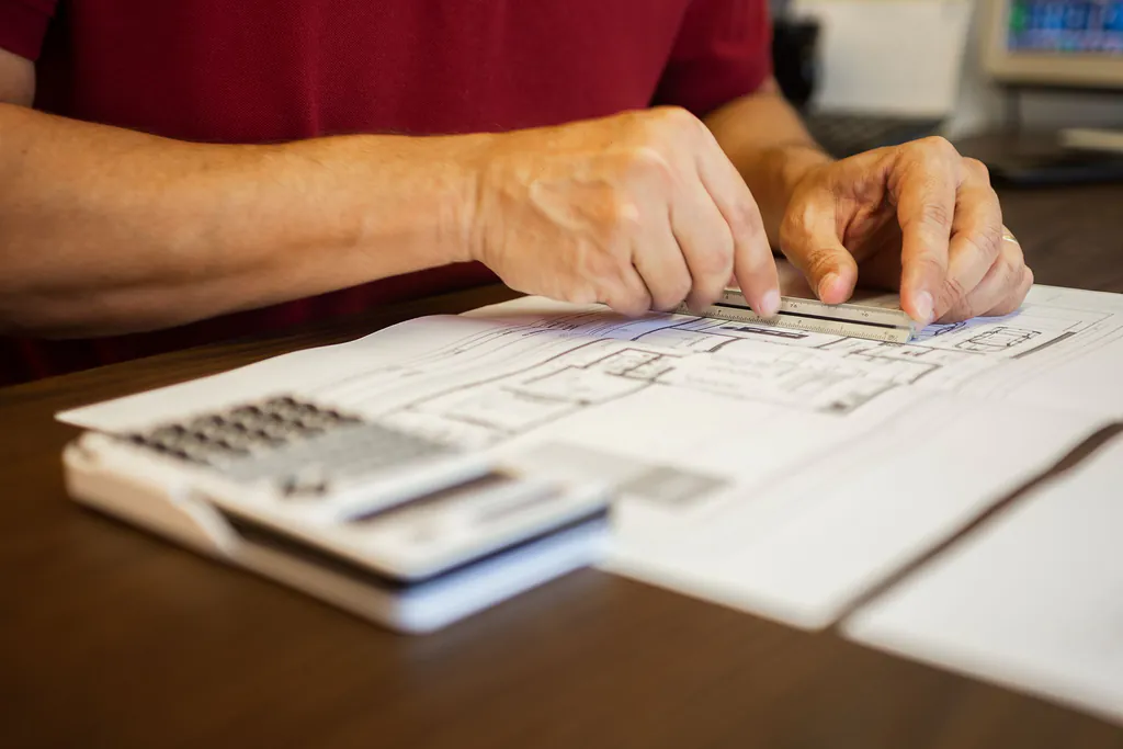 An estimator's hands holding a scale ruler against a printed plan sheet, with a calculator in the foreground, measuring runs off the drawings